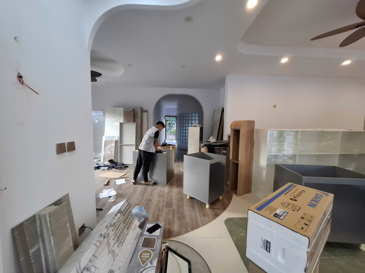 Góc nhìn từ cửa vòm vào khu vực thợ đang lắp tủ, sàn còn vật tư và bao bì bảo vệ.