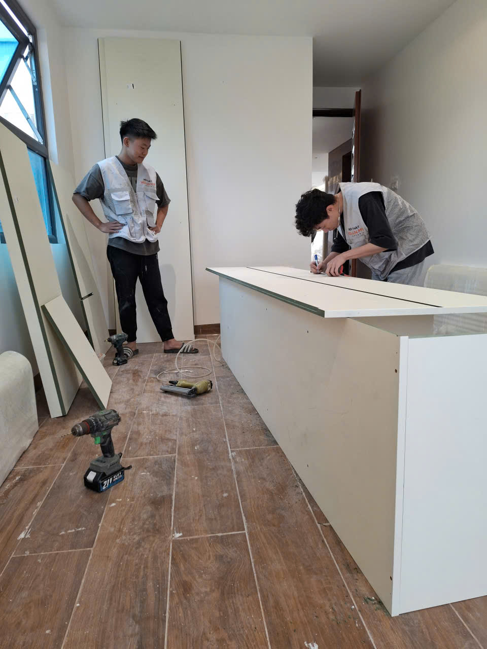Hai thợ đang căn chỉnh một thùng tủ trắng, dụng cụ lắp đặt đặt trên sàn bên cạnh.