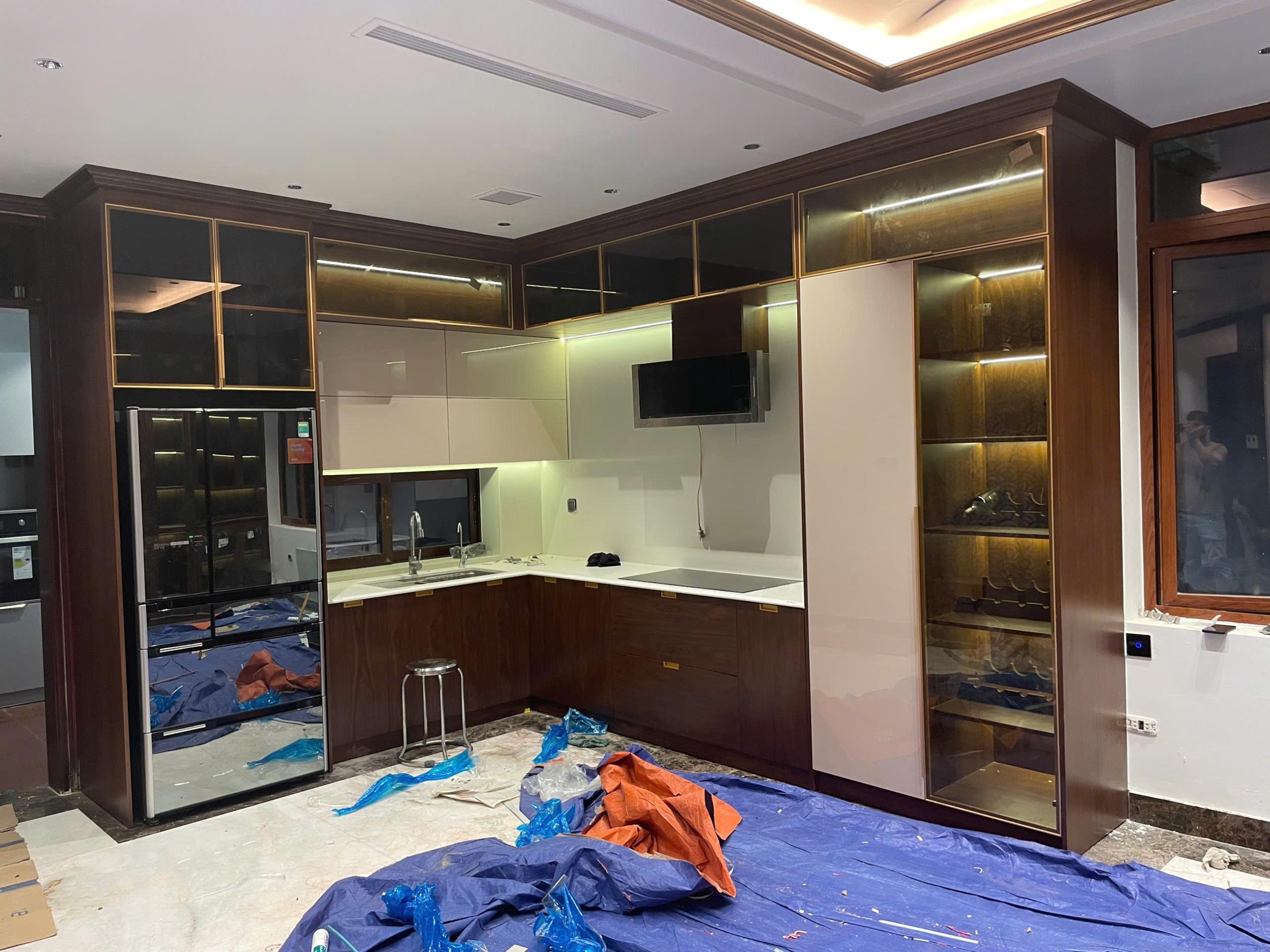 Hệ tủ bếp chữ L phối trắng và vân gỗ, kết hợp tủ kính có đèn; sàn còn phủ bạt bảo vệ.