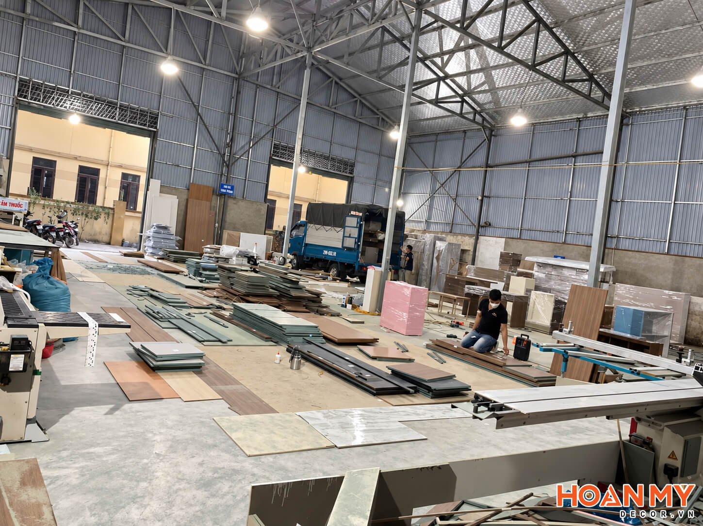 Thợ làm việc bên các bàn gia công và những chồng tấm ván trong nhà xưởng mái cao.