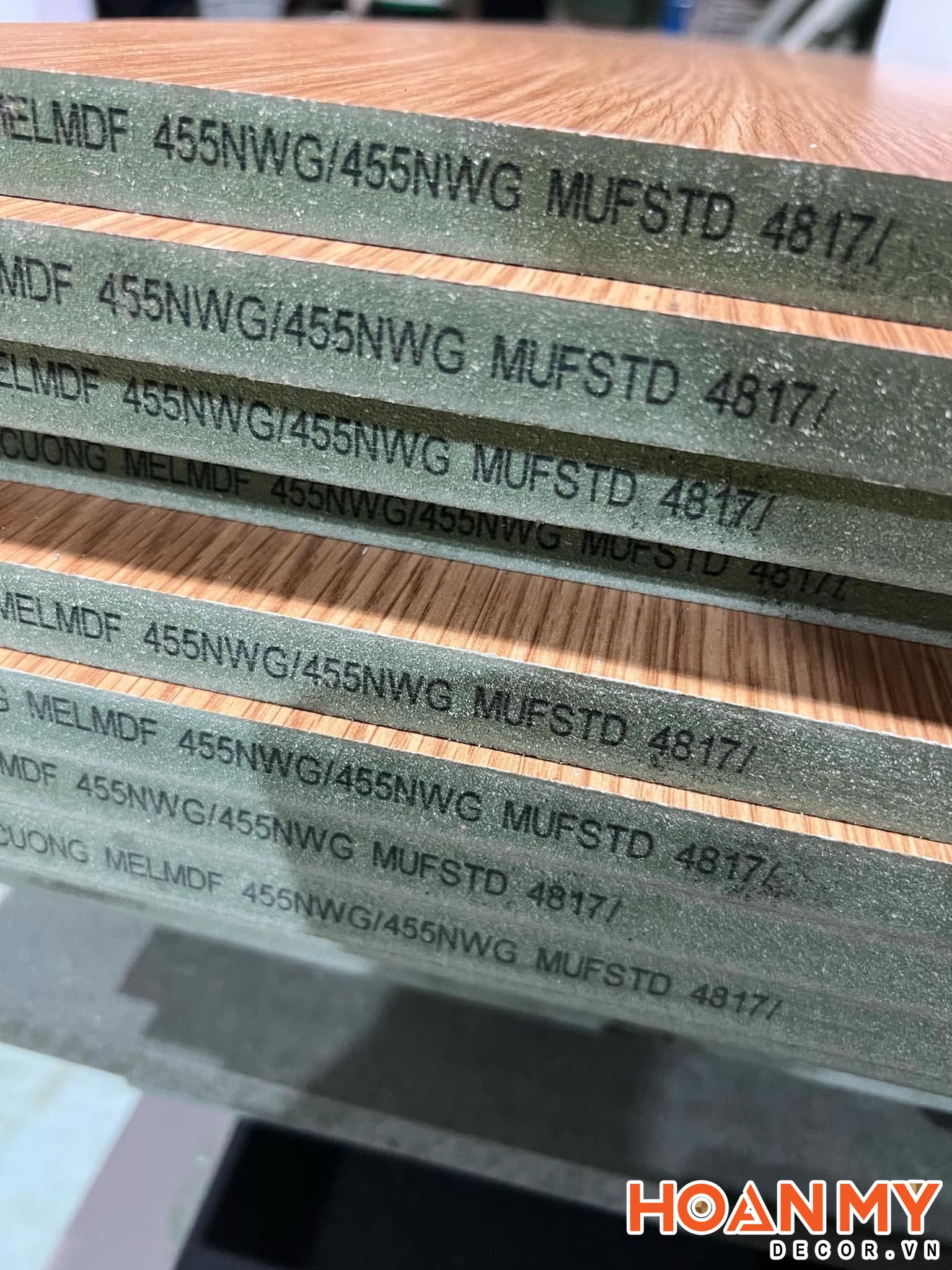 Cận cảnh chồng ván An Cường phủ melamine vân gỗ, cạnh tấm in mã MELMDF 455NWG và lộ lớp lõi MDF màu xanh.