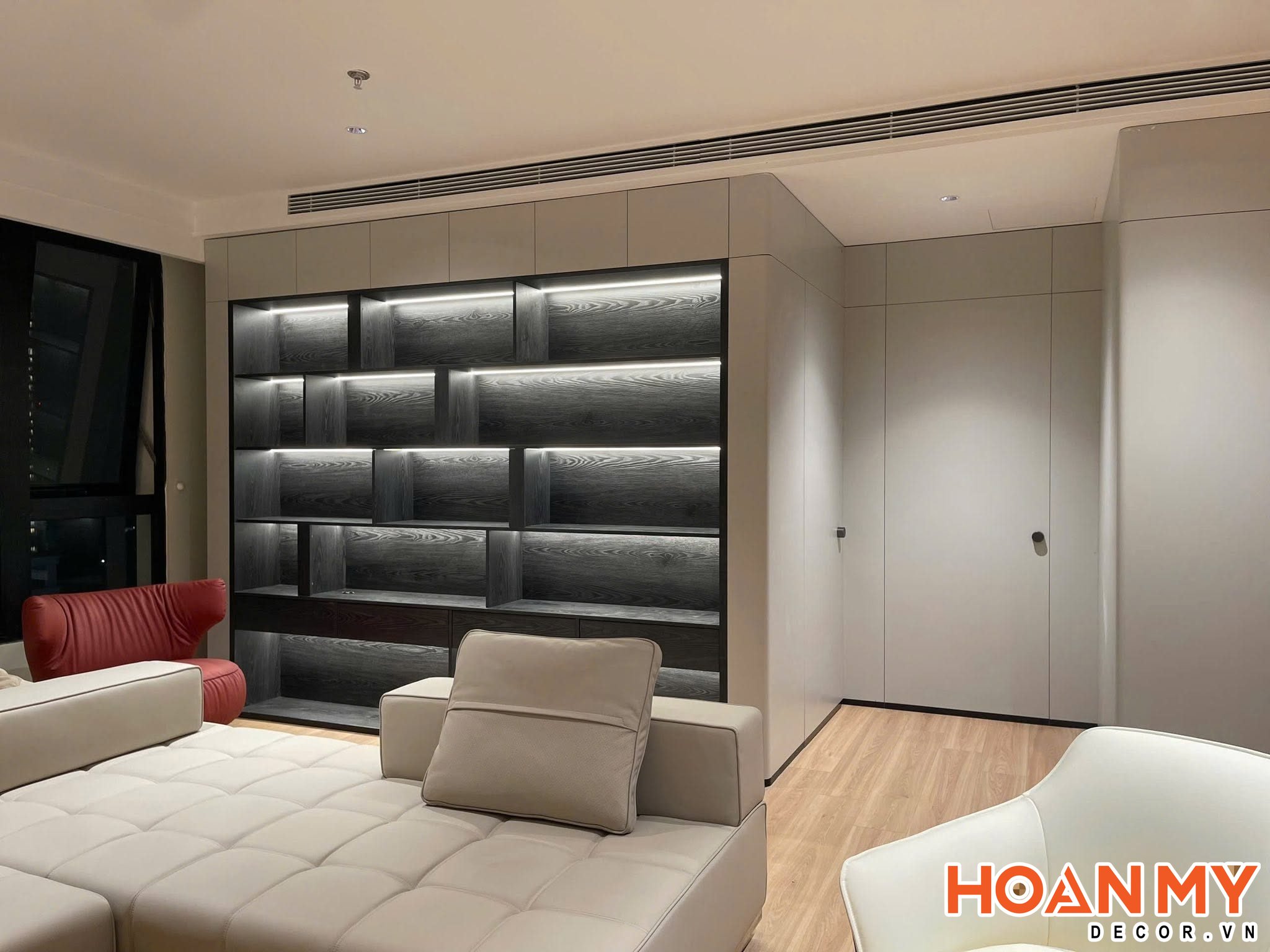 Phòng khách với tủ trang trí kịch trần khung đen nền vân sồi tối, đèn LED chạy dọc từng đợt, liền kề hệ tủ cánh trơn màu kem.