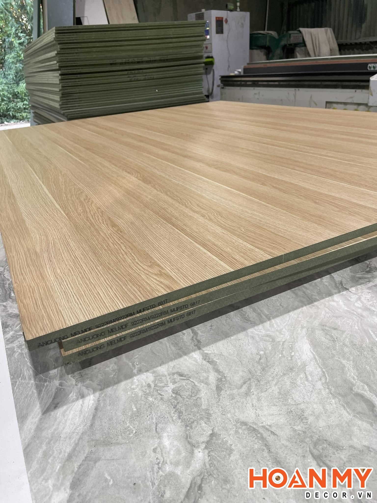 Tấm ván MDF An Cường phủ melamine vân sồi sáng tại xưởng, cạnh tấm in mã MELMDF và lộ rõ lớp lõi màu xanh.