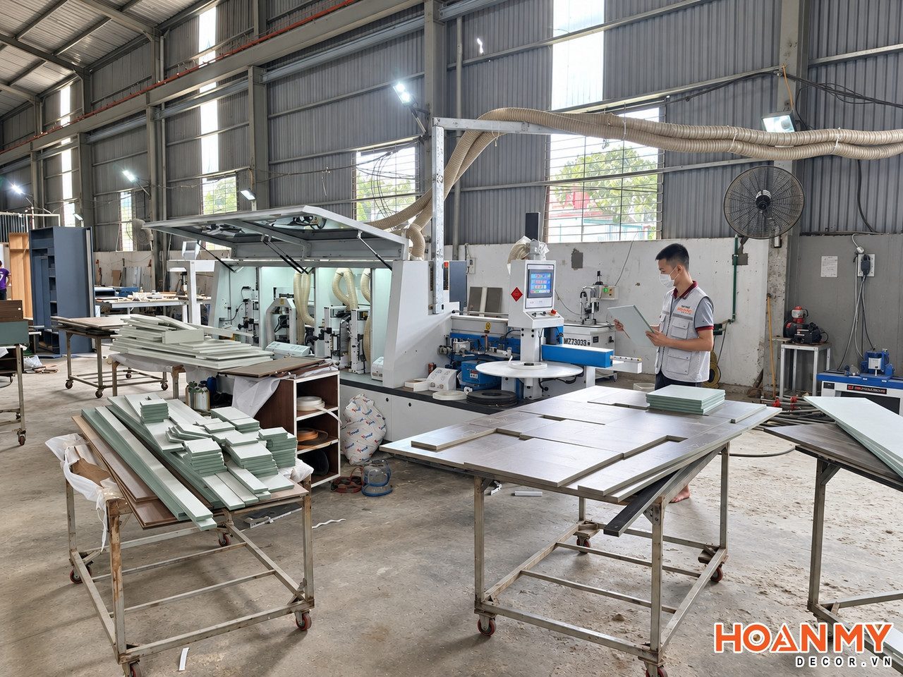 Thợ Hoàn Mỹ Decor kiểm tra tấm chi tiết bên cạnh máy dán cạnh, phía trước là các xe đẩy chở chi tiết ván đã cắt.